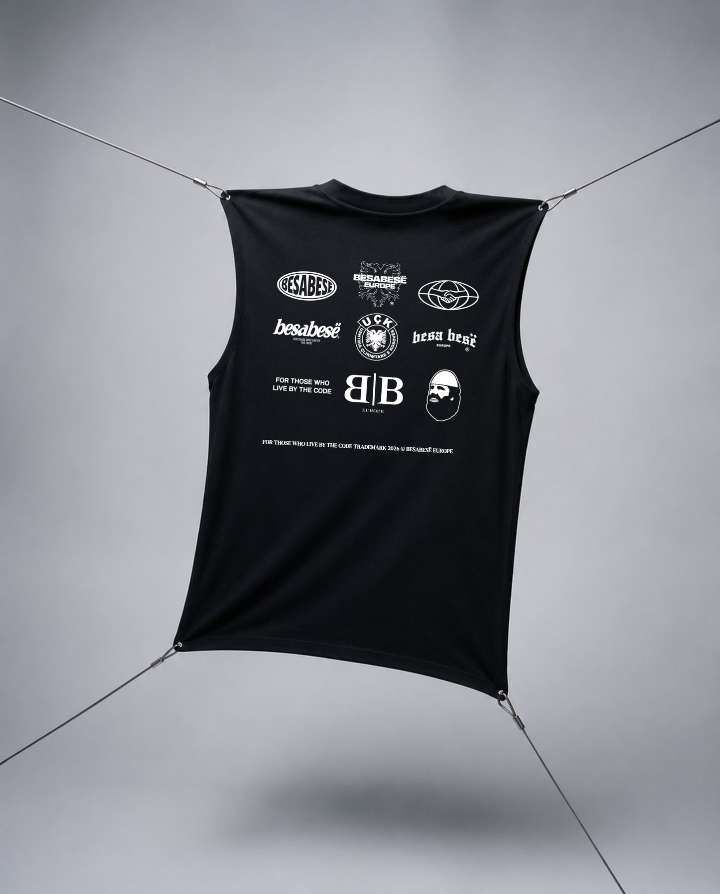A black sleeveless crop top with a white graphic print featuring multiple logos and text, suspended by thin cables against a gray studio background.