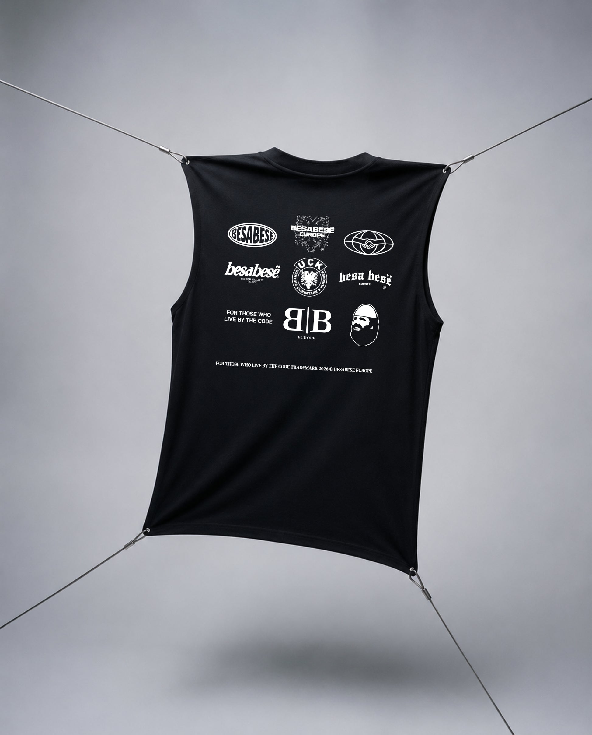 A black sleeveless crop top with a white graphic print featuring multiple logos and text, suspended by thin cables against a gray studio background.