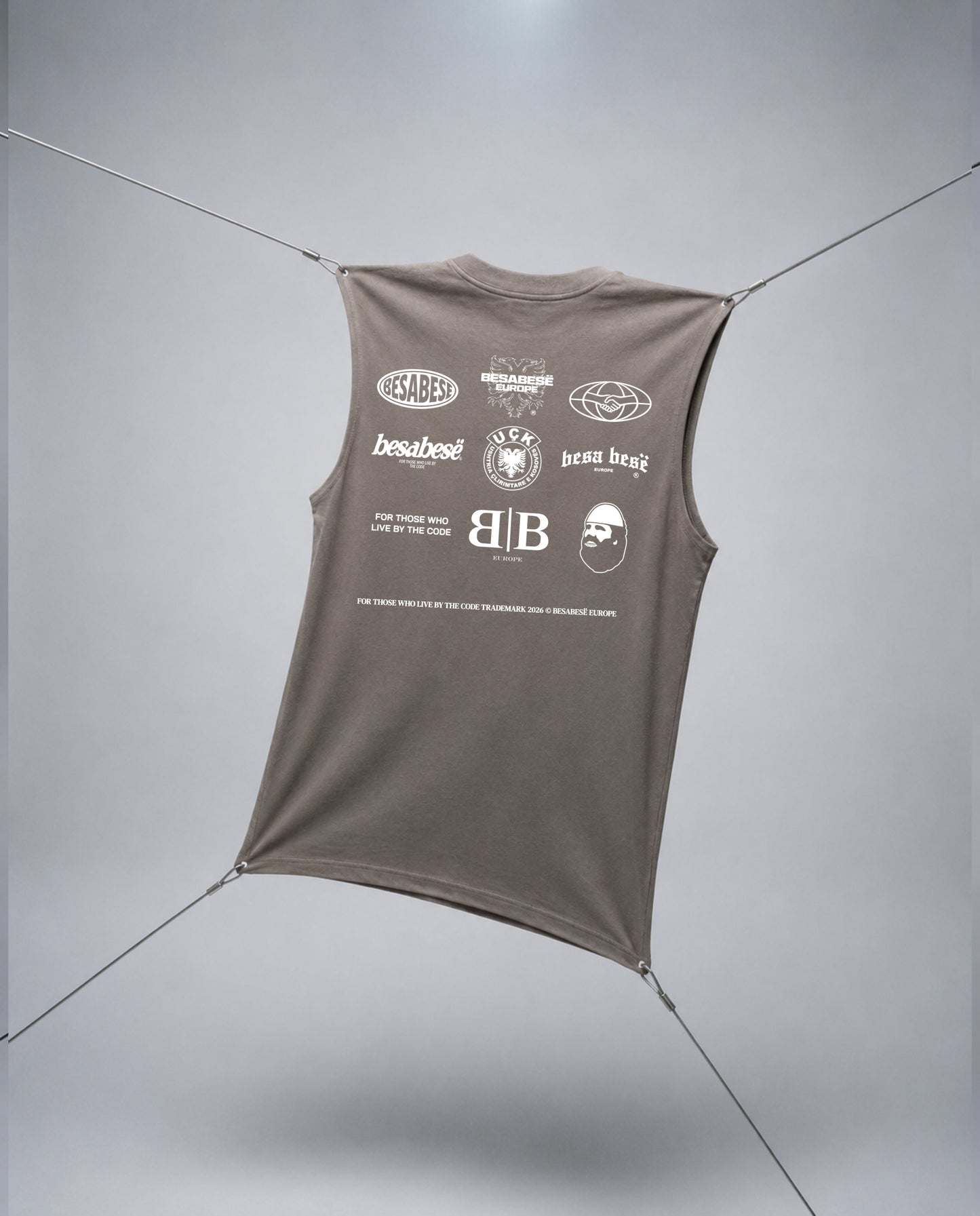A sleeveless taupe-brown t-shirt with a white graphic print on the back, suspended by thin cables against a light gray studio background.