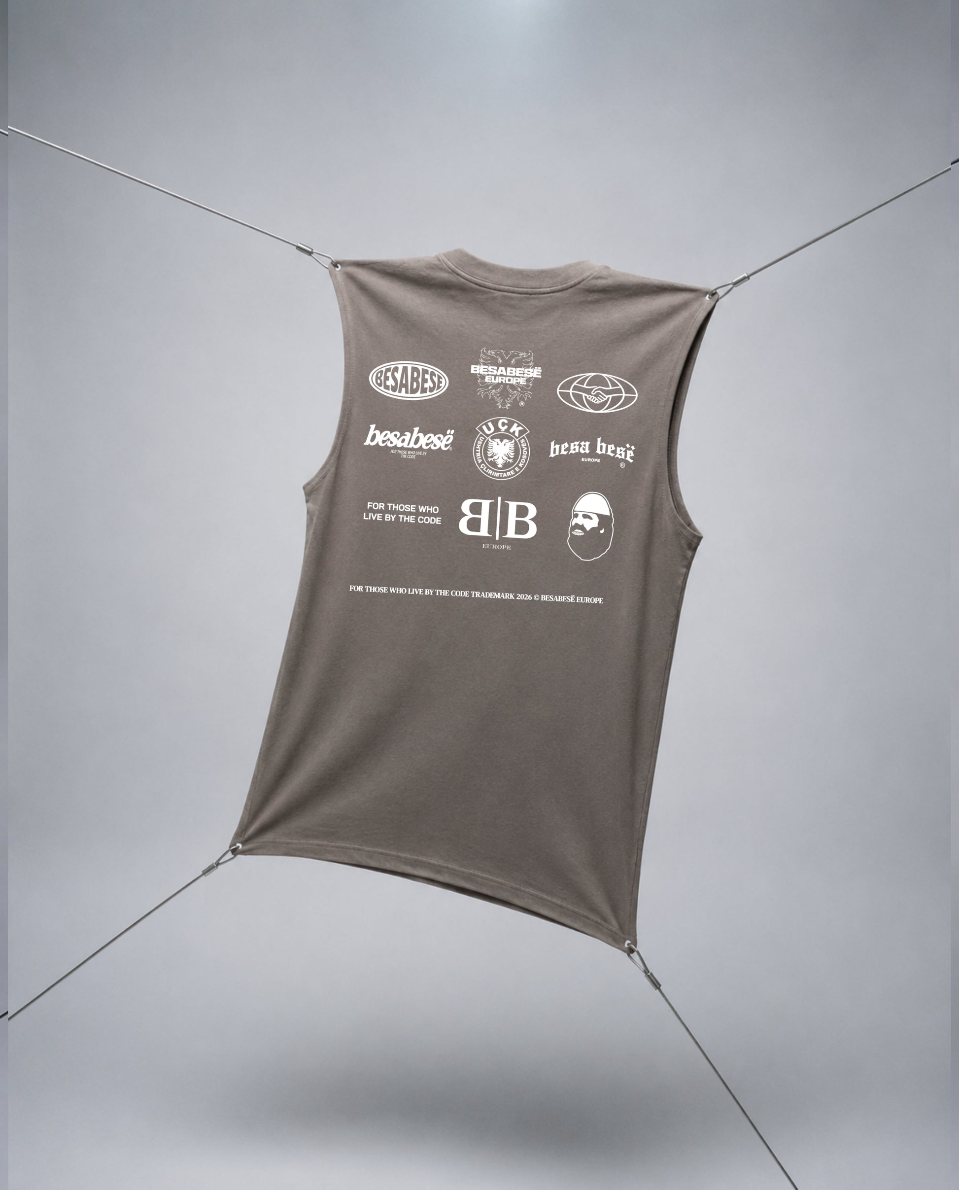 A sleeveless taupe-brown t-shirt with a white graphic print on the back, suspended by thin cables against a light gray studio background.