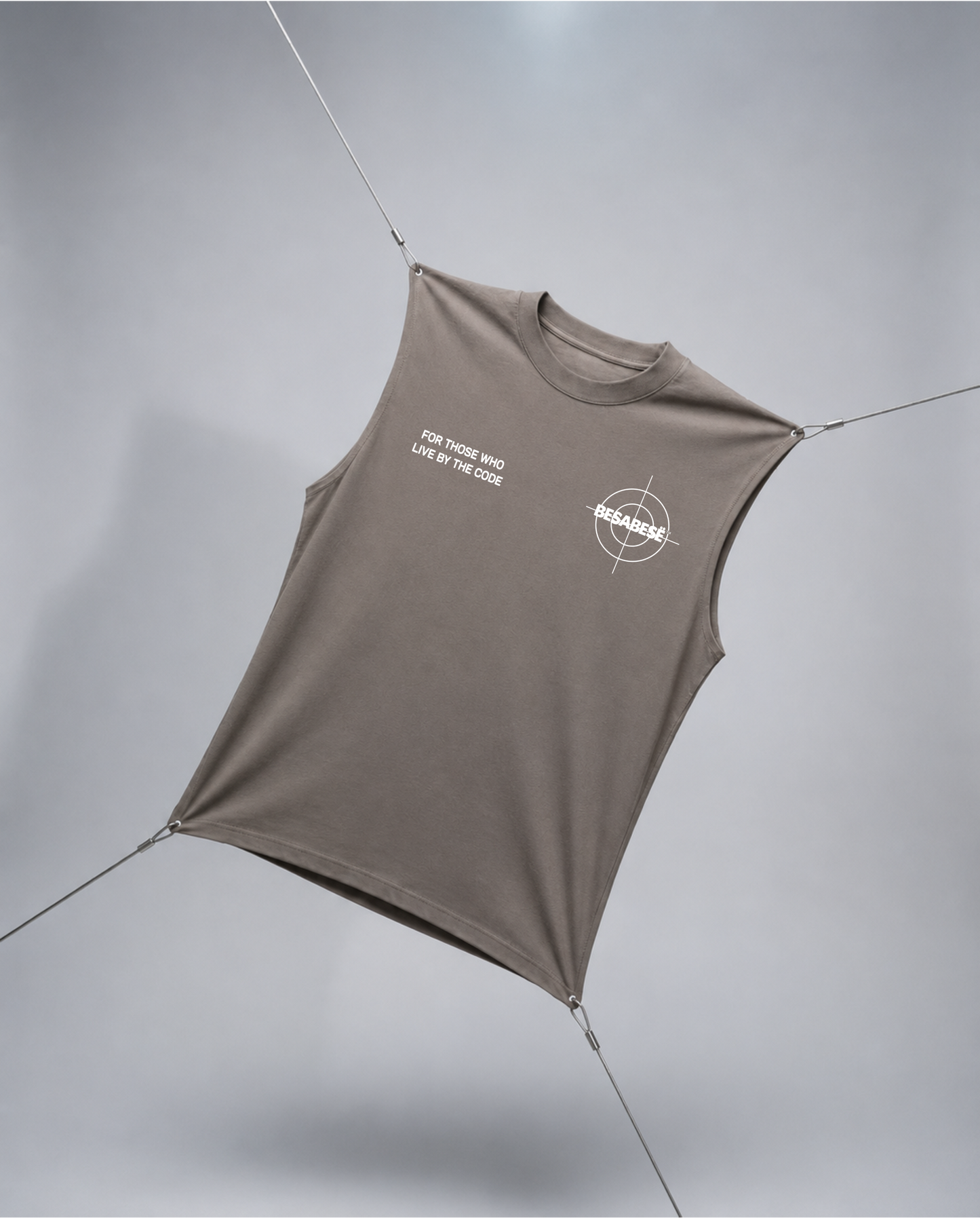 A sleeveless fitted tank top in taupe-brown with white printed graphics on the chest, suspended by thin cords against a light gray studio background.