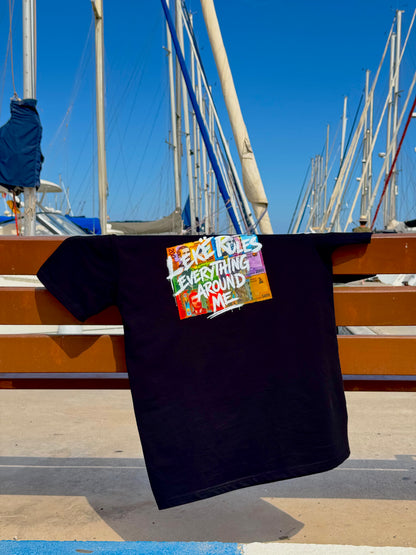 A black t-shirt hanging from a wooden railing at a marina, featuring a colorful graphic print that reads 'LEKE RULES EVERYTHING AROUND ME'.